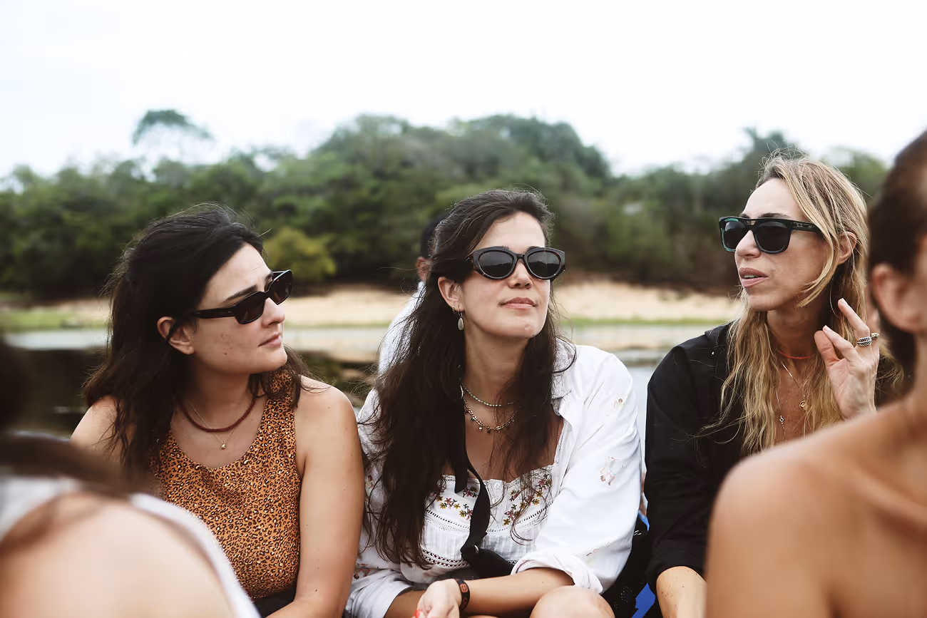 Three women wearing sunglasses sitting outdoors near water and greenery, engaged in conversation.