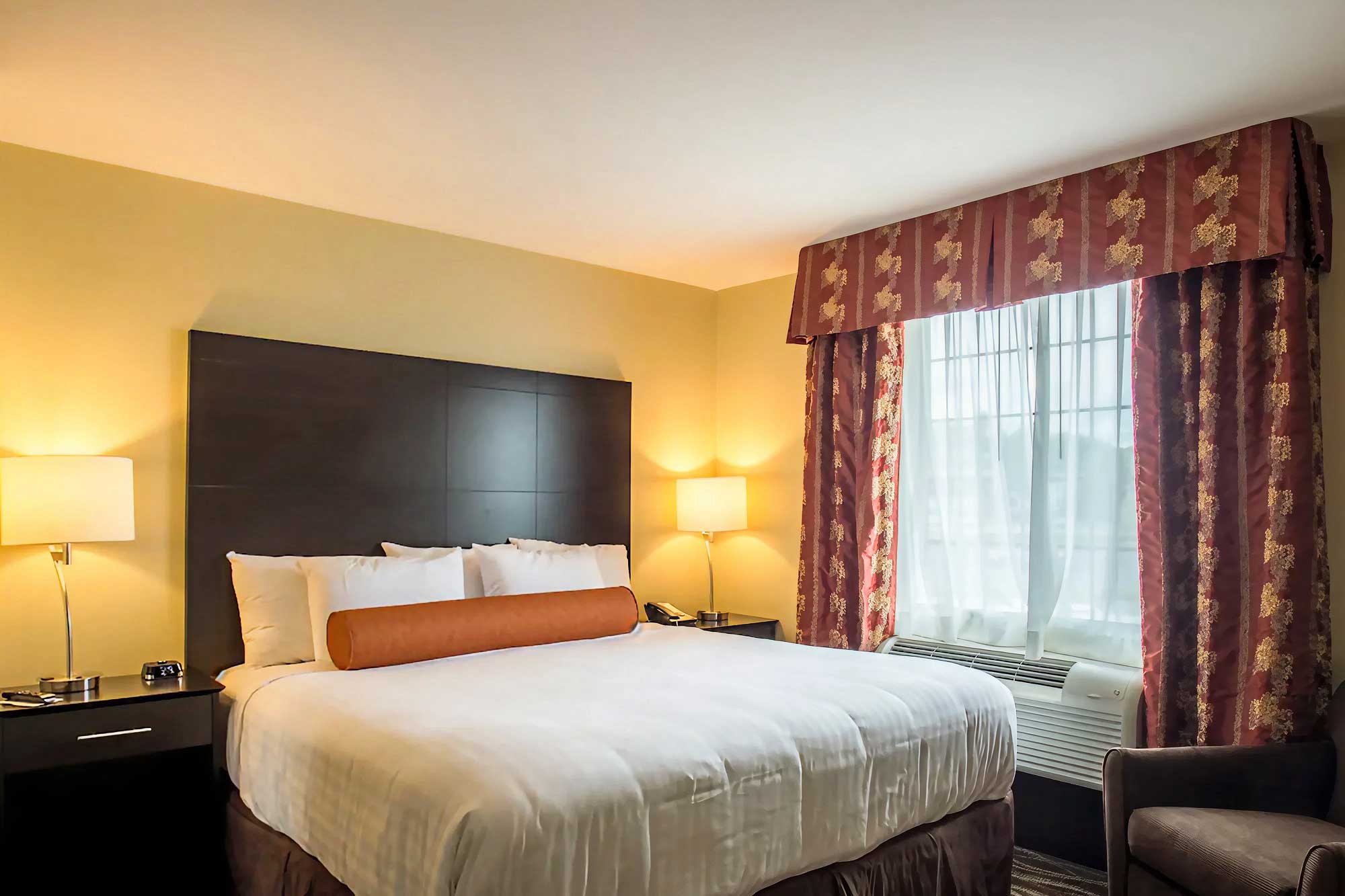 Hotel king room with a single king bed, dark wood headboard, bedside tables with lamps, upholstered chair, and window with patterned curtains.