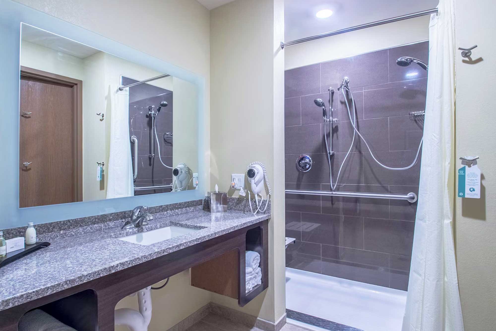 Modern accessible bathroom with granite countertop, mounted hairdryer, large mirror, and walk-in shower with grab bars.