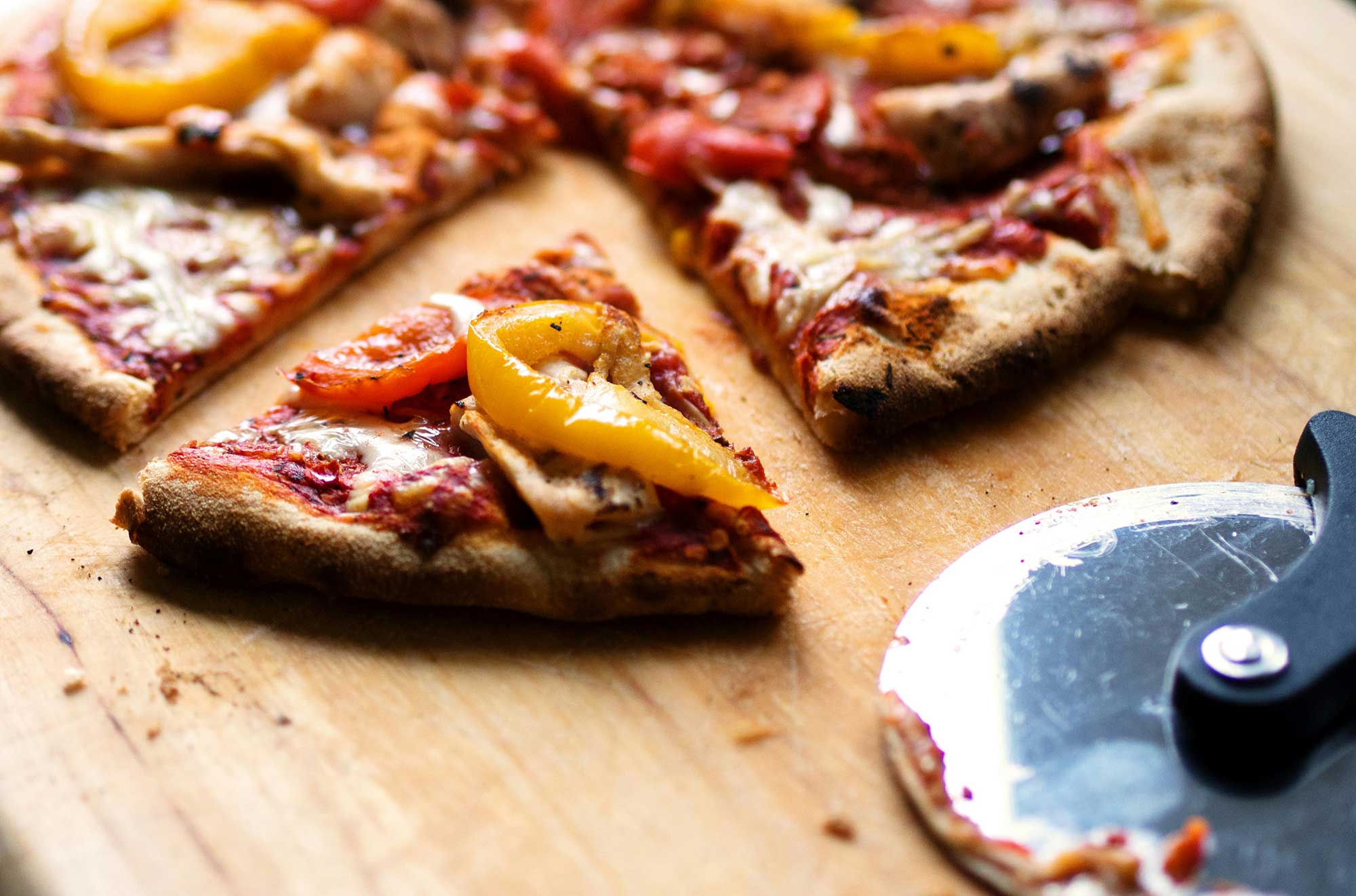 Close-up of three pizza slices topped with yellow bell peppers and grilled chicken on a wooden surface next to a metal pizza cutter.