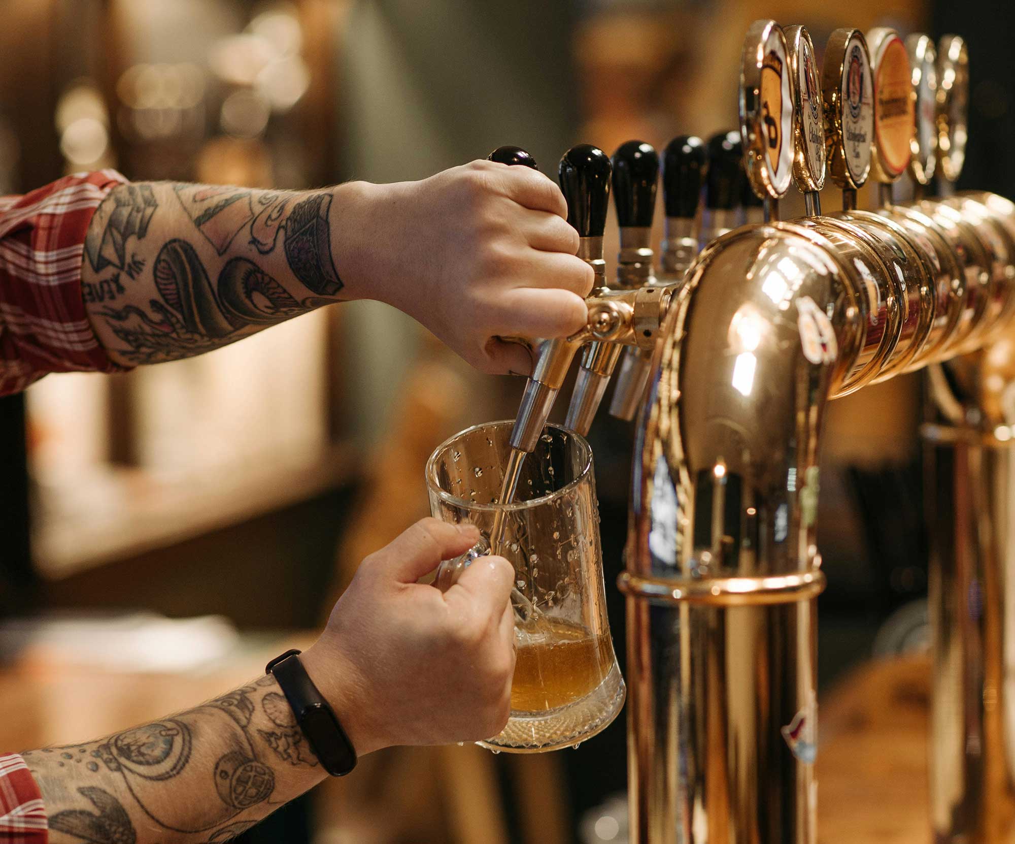 Tattooed person pouring draft beer from a tap into a glass mug at a bar.