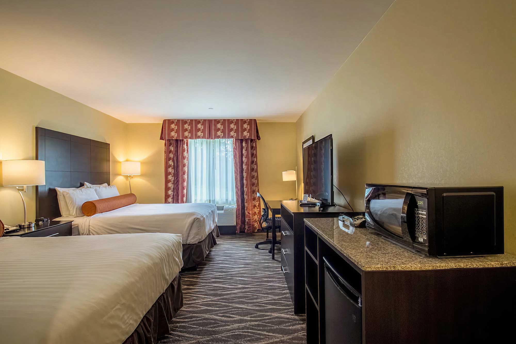 Hotel room with two queen beds, work desk and chair, flat-screen TV, microwave and mini fridge, and window with patterned curtains.