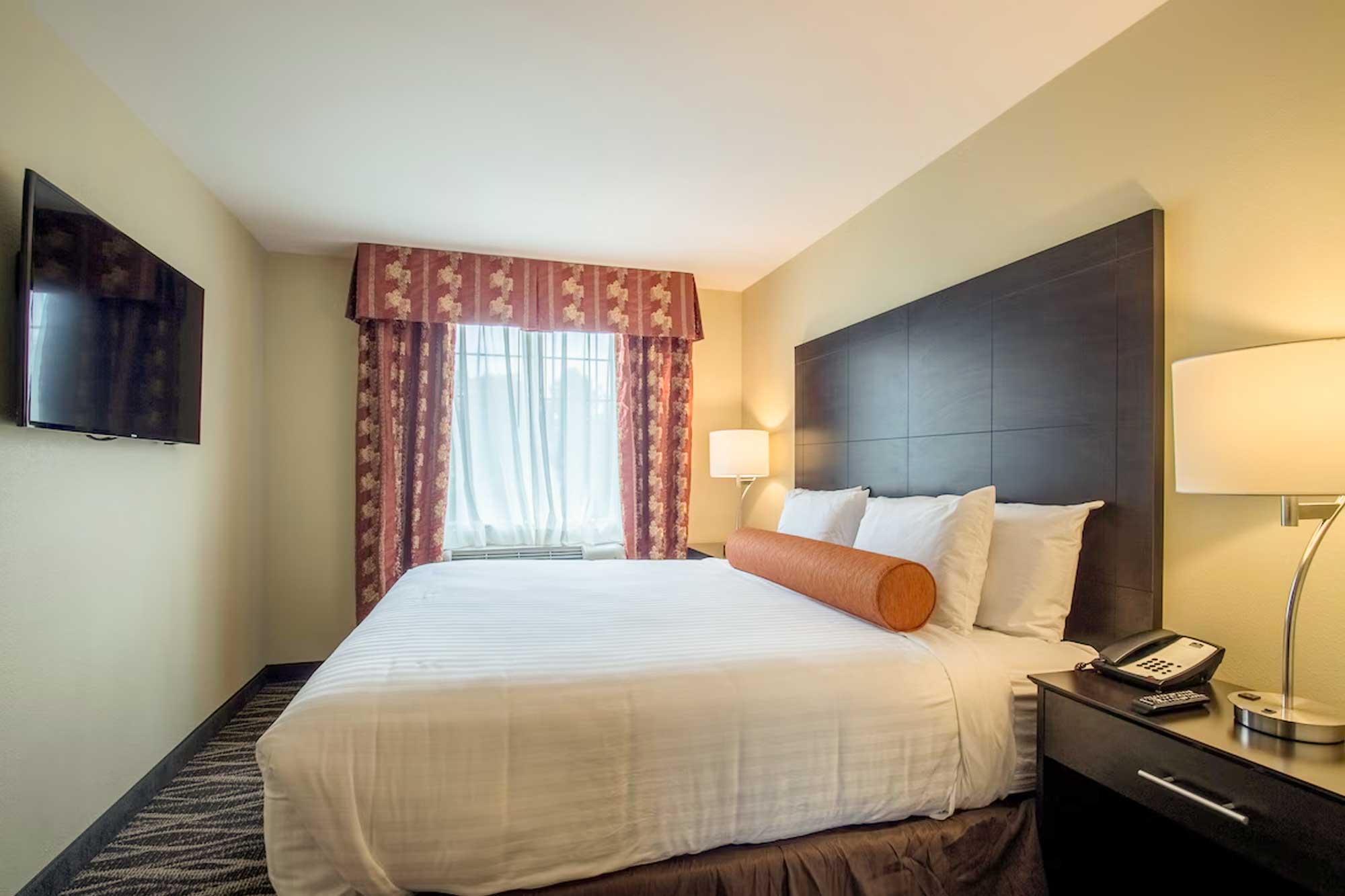 Hotel king room with a single king bed, dark wood headboard, bedside tables with lamps, upholstered chair, and window with patterned curtains.