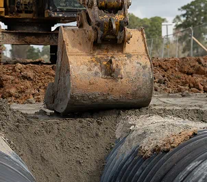 Excavator bucket digging soil near large black drainage pipes in a construction site.