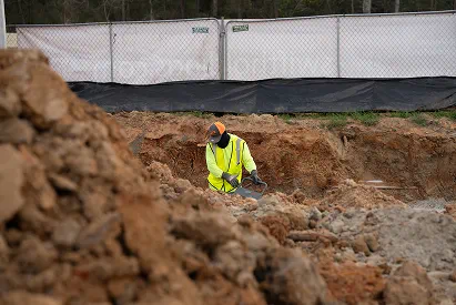 Construction worker in a yellow safety vest working in a deep excavation site with dirt piles around.