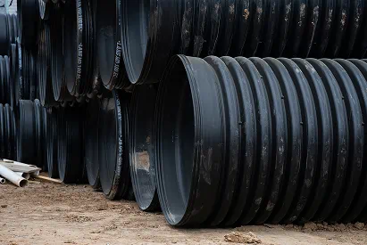 Stack of large black corrugated drainage pipes on a dirt ground.