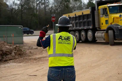 Construction worker in a hard hat and reflective vest signaling near a yellow dump truck on a dirt road.