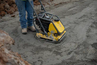 Person operating a yellow soil compactor on a construction site.