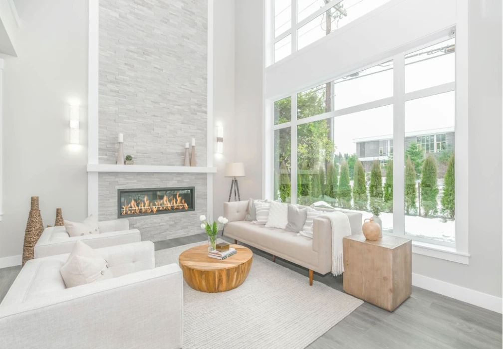 A bright, modern living room interior featuring a stone fireplace and large windows overlooking greenery.