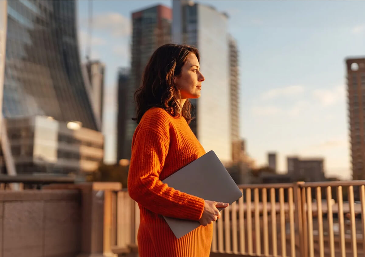 Woman in an orange sweater holding a laptop, standing on a balcony with city buildings in the background at sunset.