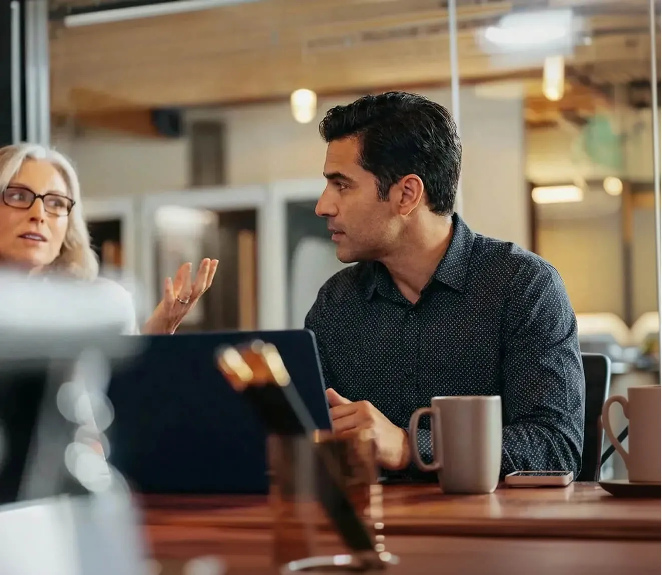 Two colleagues engaged in a discussion at a table with mugs and a tablet in an office setting.