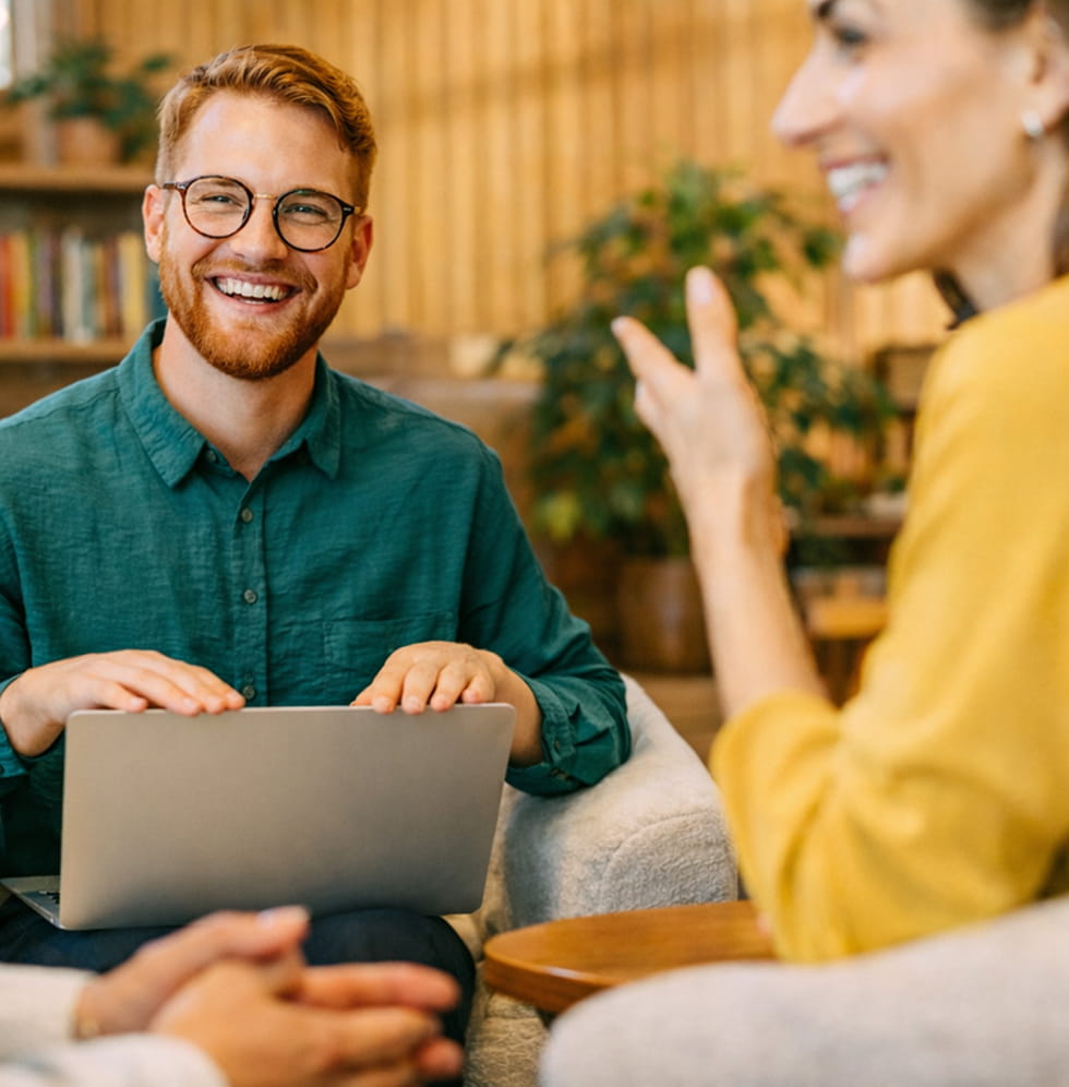 Smiling man with glasses in a green shirt holding a laptop, engaged in conversation with two people.
