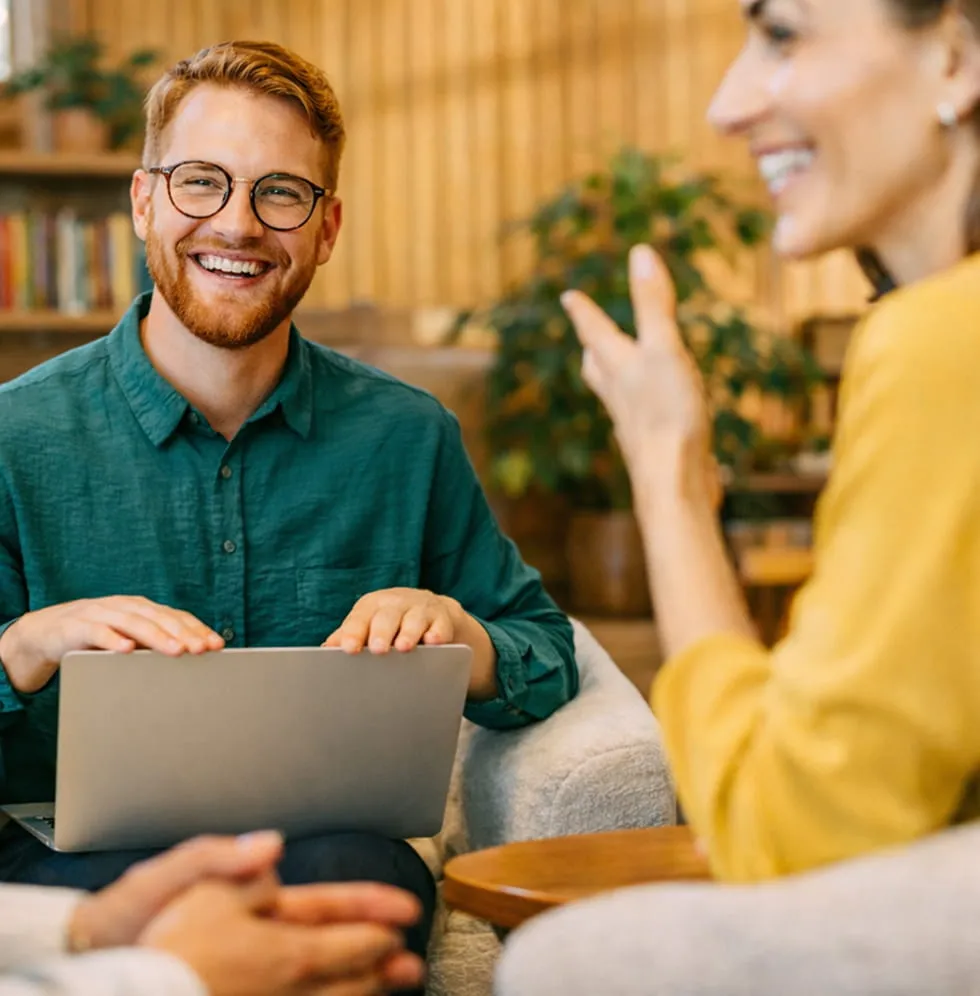 Smiling man with glasses in a green shirt holding a laptop, engaged in conversation with two people.