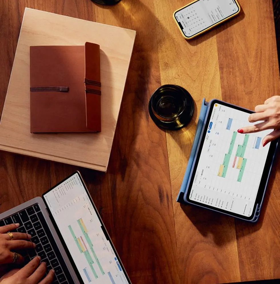 Overhead view of a wooden table with a brown notebook, a smartphone showing a calendar app, a cup, a laptop, and a tablet displaying colorful schedules, with two people interacting with the devices.
