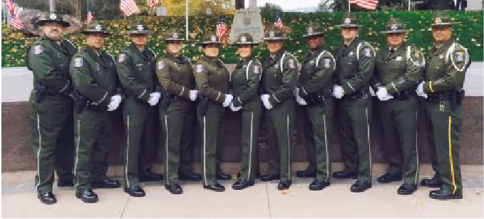 Twelve uniformed law enforcement officers standing side by side in front of a memorial with American flags.