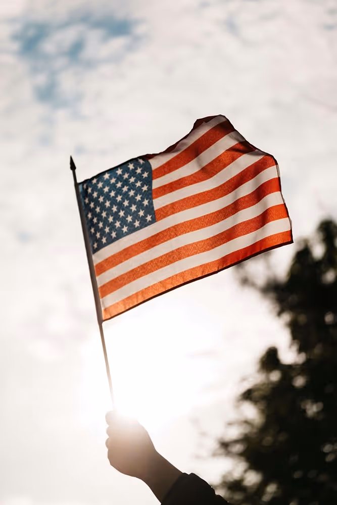 Hand holding an American flag waving against a bright sky with sun shining through.