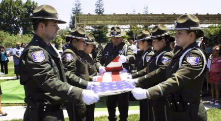 Uniformed officers in ceremonial hats folding an American flag during a memorial service outdoors.
