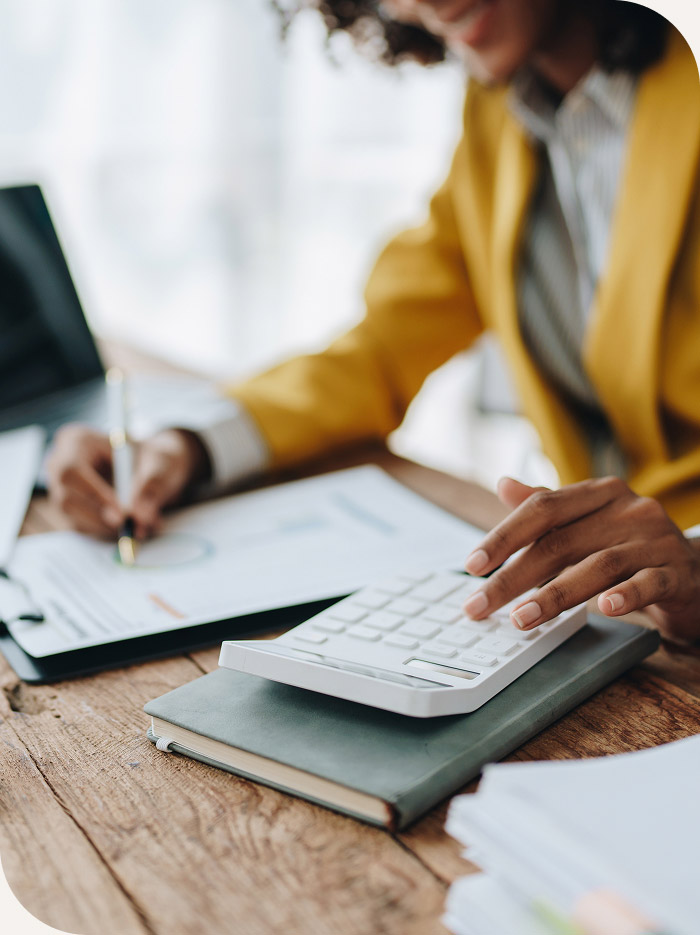 Person in a mustard jacket using a calculator and writing on a clipboard with documents on a wooden desk.