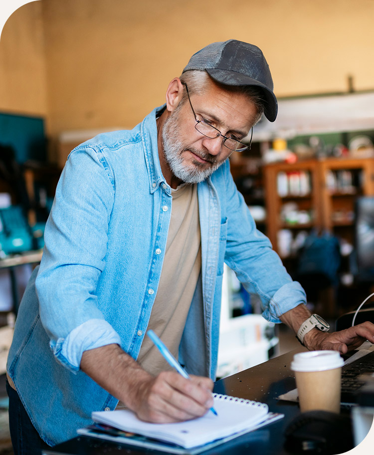 Man wearing glasses and a cap writing in a notebook on a desk with a coffee cup nearby.