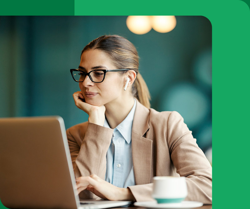 Woman wearing glasses and a beige jacket working on a laptop with wireless earbuds in a modern indoor setting.