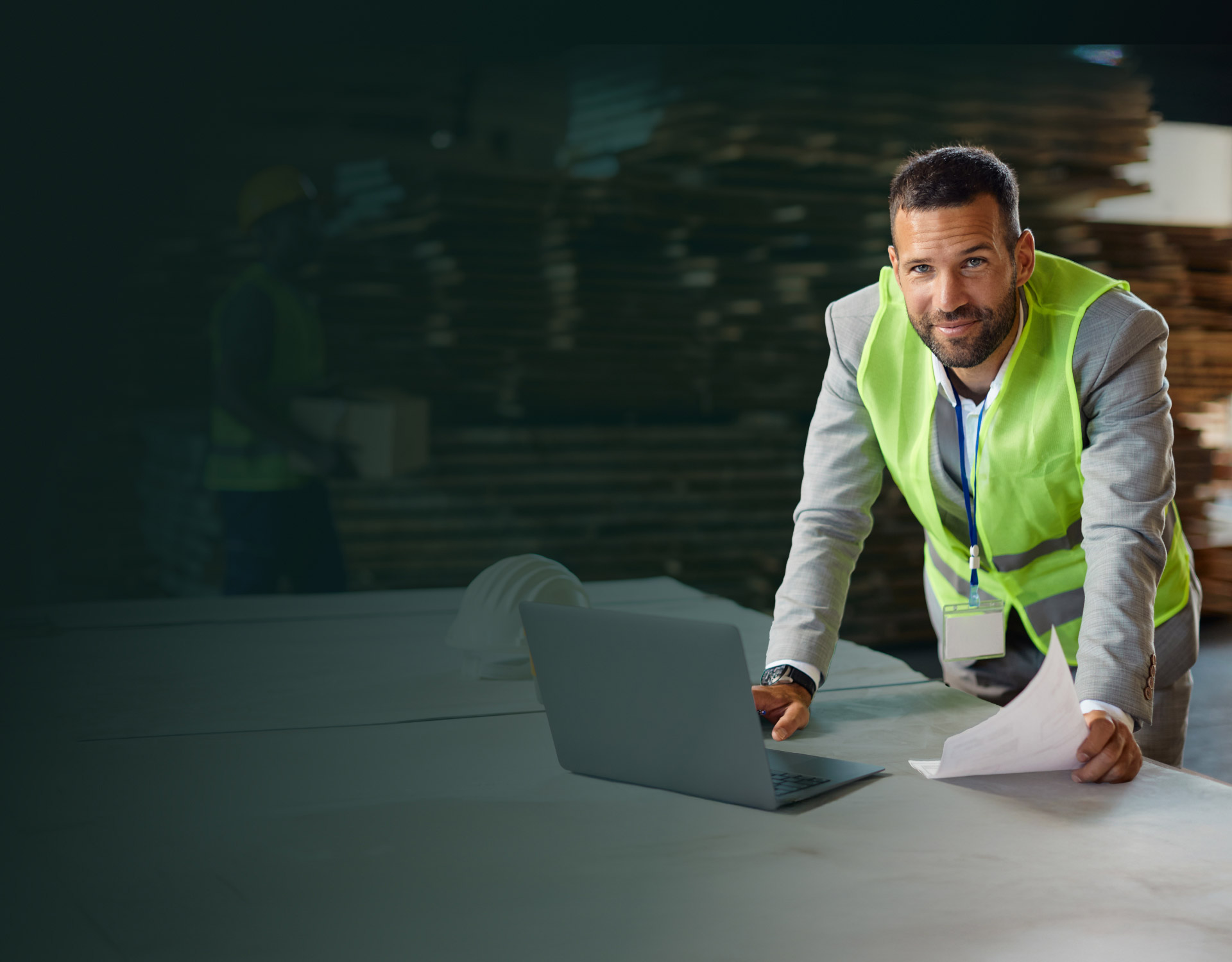Man in a gray suit and green safety vest leaning on a table with a laptop and holding documents in a warehouse with stacks of wood.