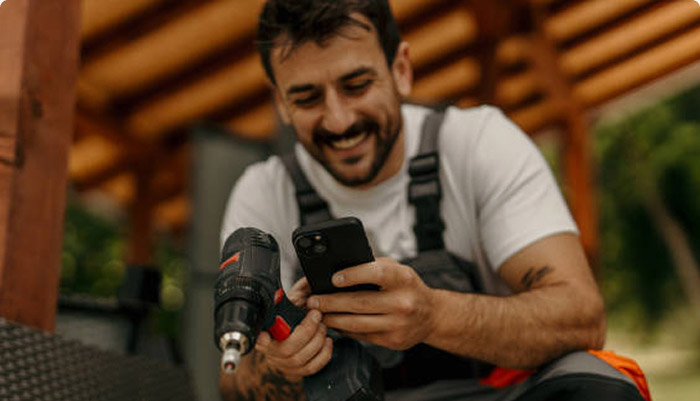 Smiling man in work overalls holding a power drill and looking at his smartphone.