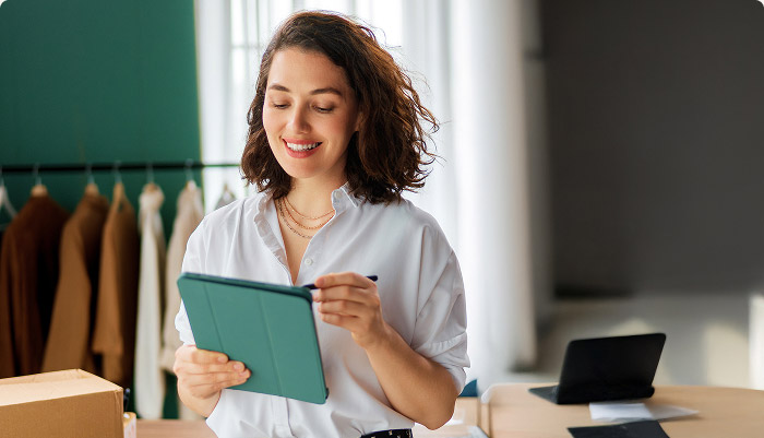 Smiling woman using a tablet with a stylus in a workspace with clothes hanging in the background.