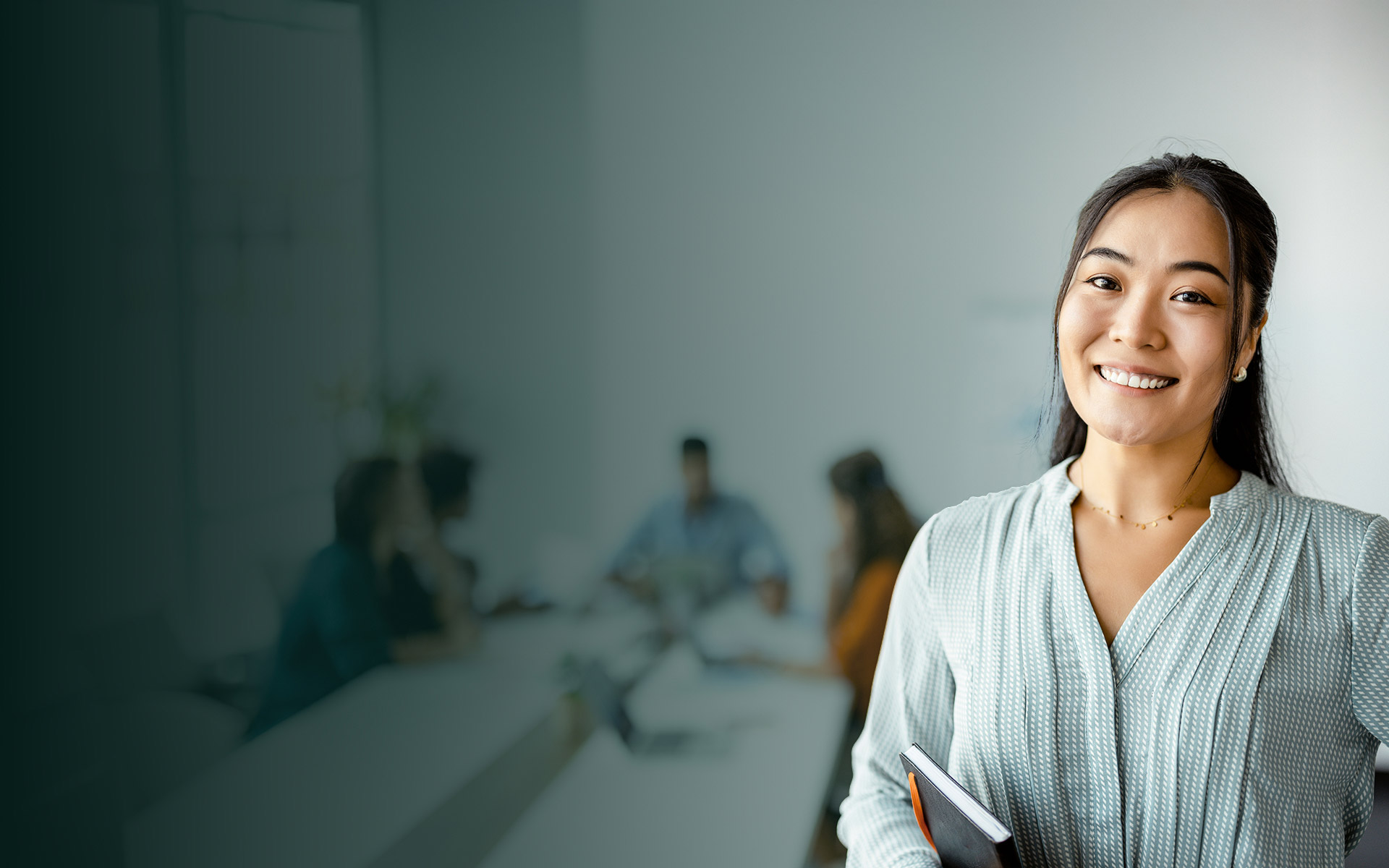 Smiling woman holding a notebook in a conference room with a blurred group meeting in the background.