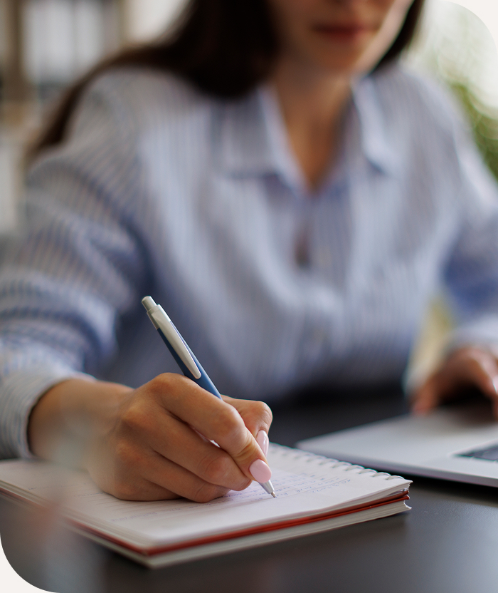 Person in a striped shirt writing in a notebook with a pen, laptop partially visible on the desk.