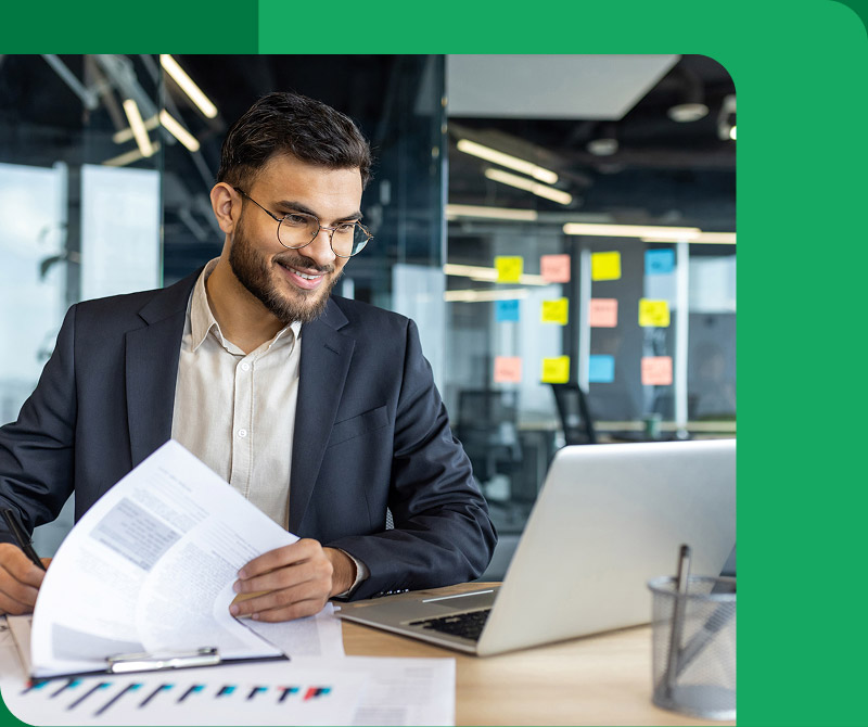 Smiling man in glasses and suit sitting at desk with papers and laptop in modern office.