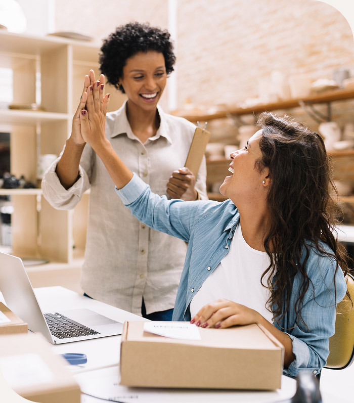 Two women smiling and giving a high-five in a workspace with a laptop and a cardboard box on the table.