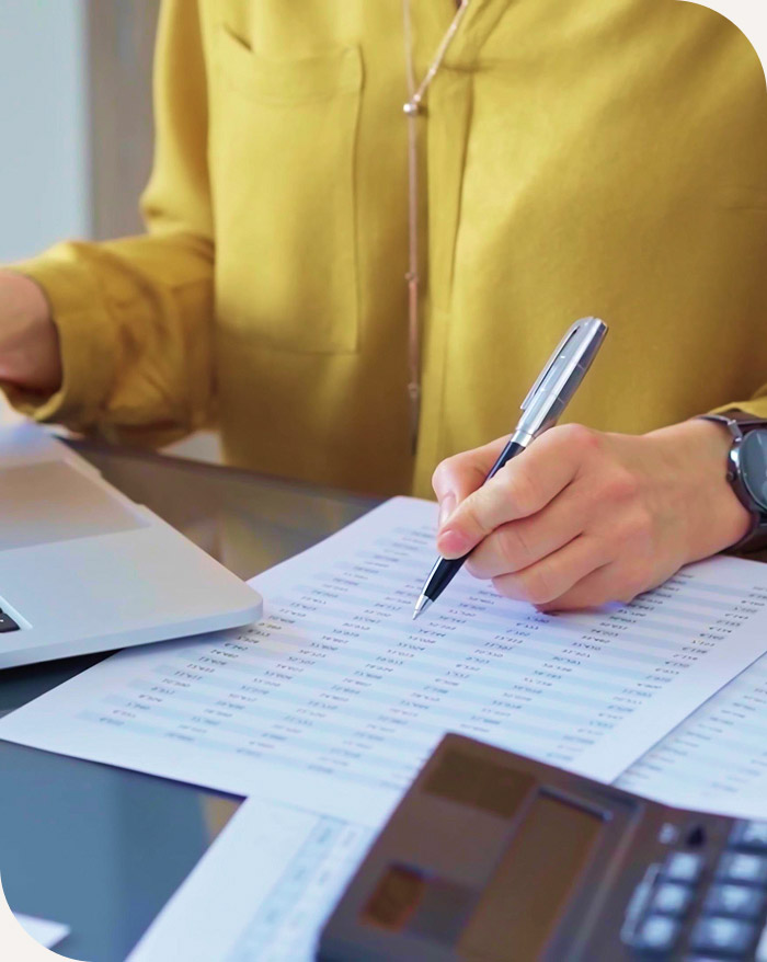 Person in yellow shirt marking numbers on a financial document next to a laptop and calculator.