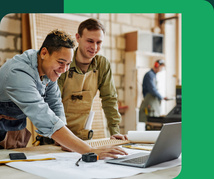 Two architects or designers smiling and reviewing plans on a laptop in a workshop.
