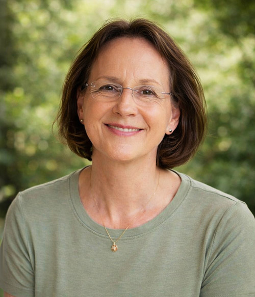 Smiling middle-aged woman with short brown hair, wearing glasses, a light green shirt, and a gold necklace, with a blurred green background.