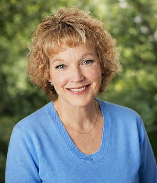 Smiling middle-aged woman with curly blonde hair wearing a light blue sweater and a gold necklace, standing outdoors with blurred green foliage background.