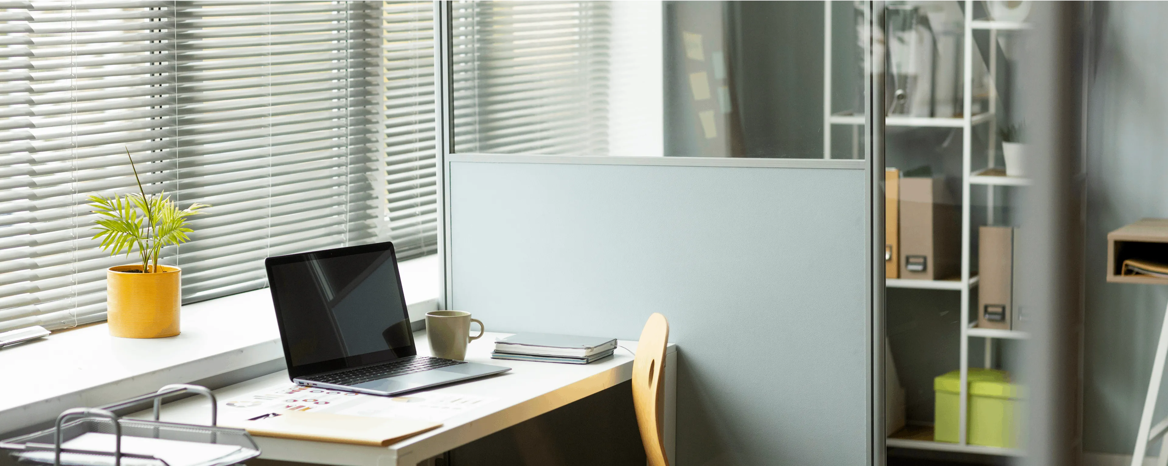 Modern office desk with a laptop, coffee mug, notebooks, and a yellow potted plant next to a window with blinds.