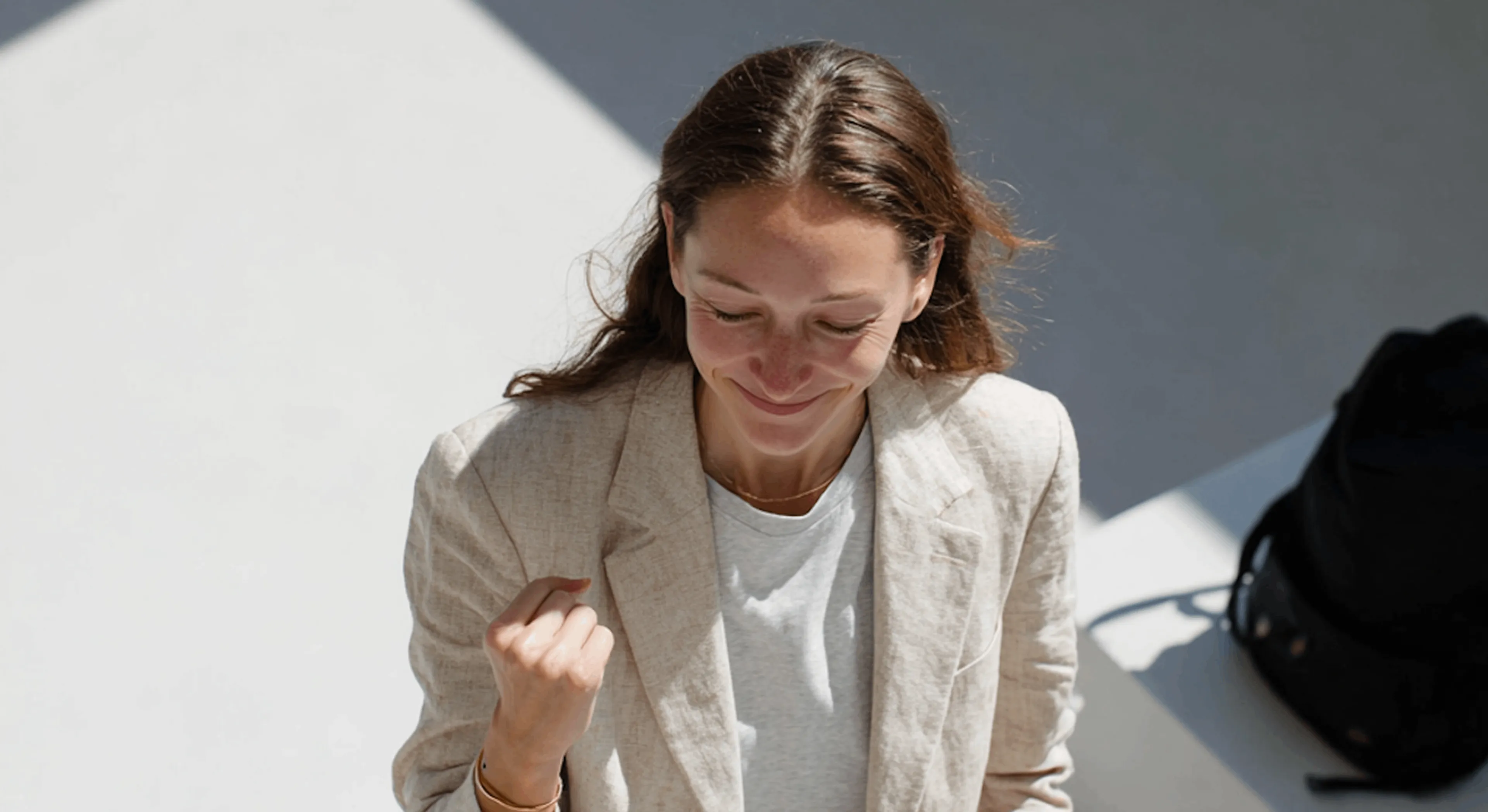Smiling young woman with brown hair wearing a beige blazer and white shirt, clenching her fist in a gesture of excitement.