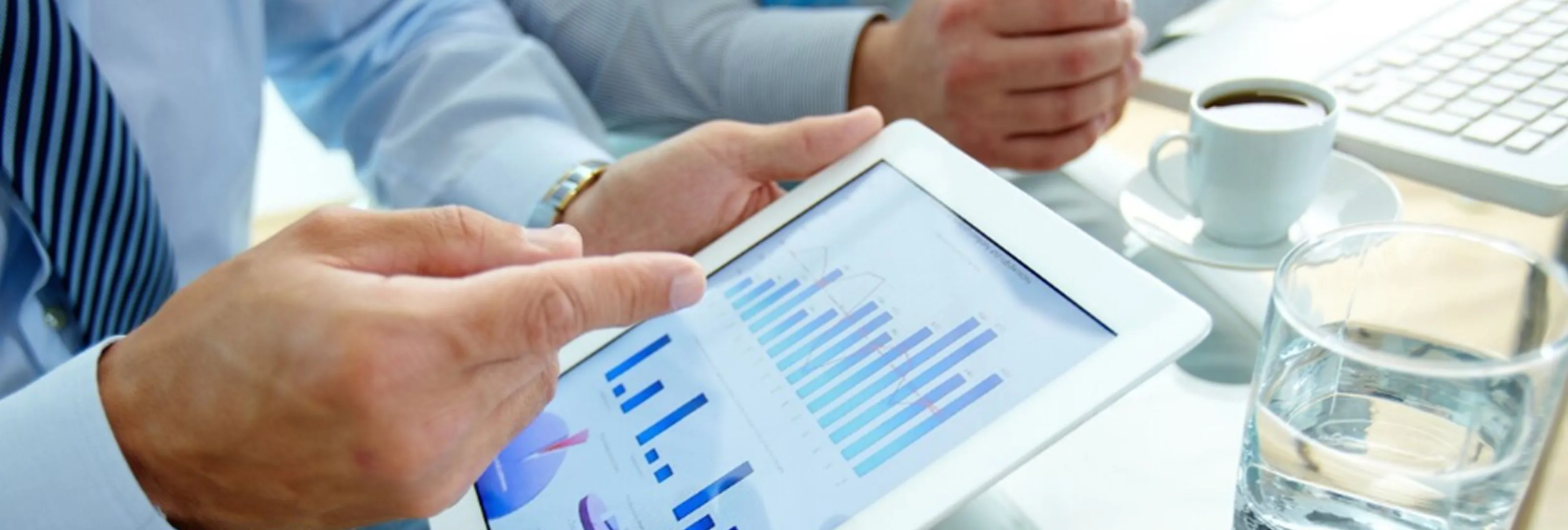 Two people in business attire reviewing charts and graphs on a tablet, with a cup of coffee and a glass of water nearby.