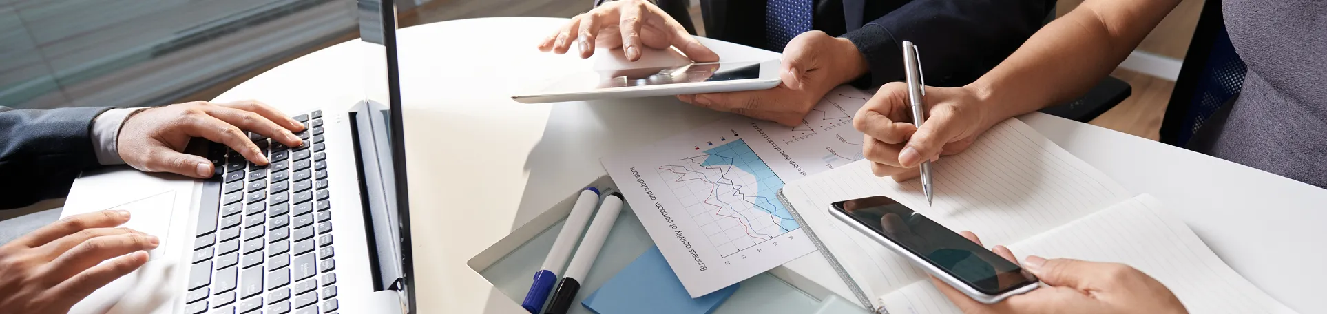 Three people working together at a round table with laptop, tablet, financial charts, pens, notebook, and smartphone.