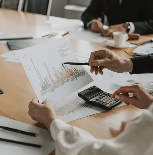 Two people reviewing a printed bar chart with a pen and calculator on a wooden table.