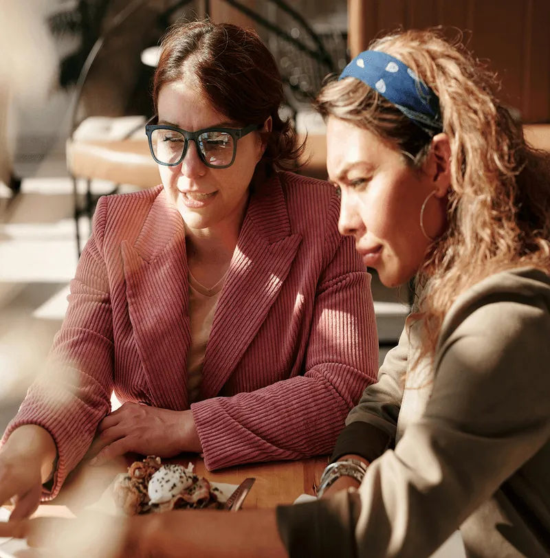 Two women sitting and looking at a smartphone on a wooden table with a plate of food.