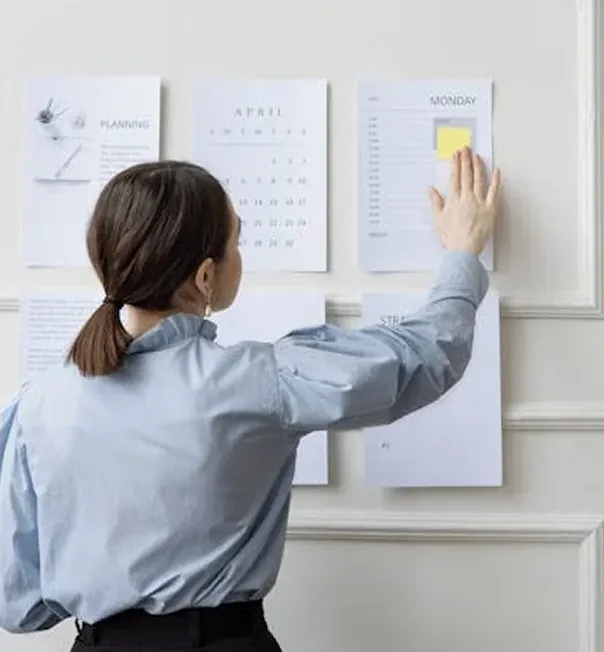 Woman in a blue shirt looks at and touches a wall calendar with planning sheets, including a calendar for April and a Monday schedule.