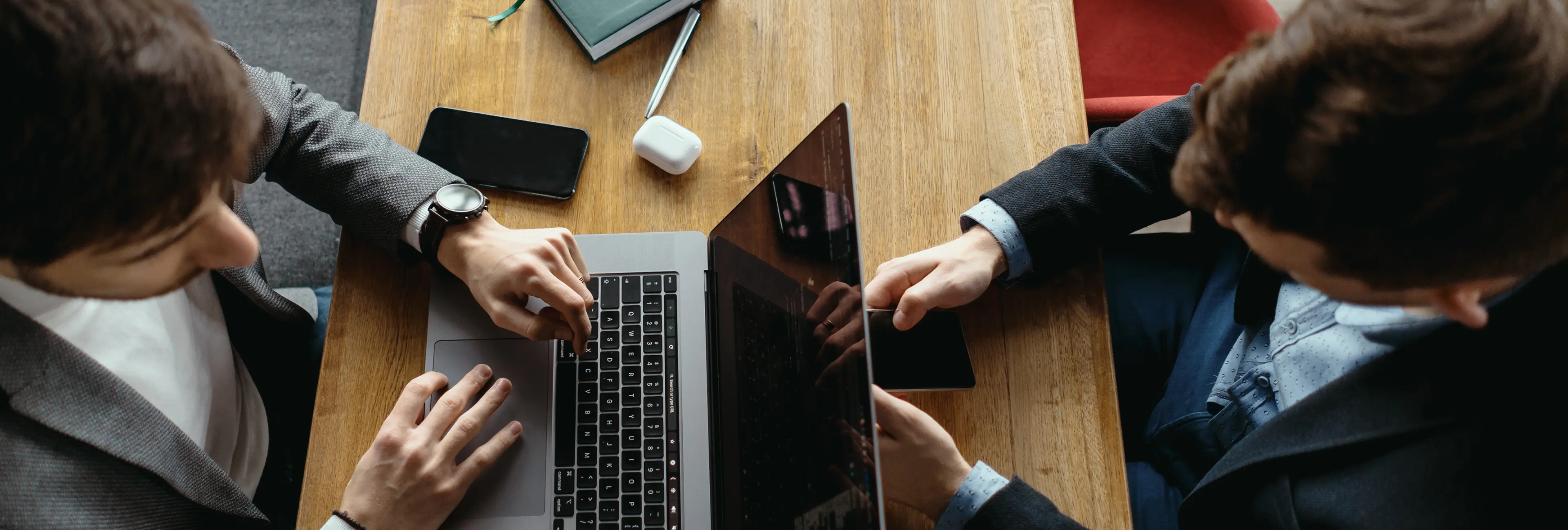 Two people working together on a laptop at a wooden table with a phone, earbuds case, and a notebook nearby.