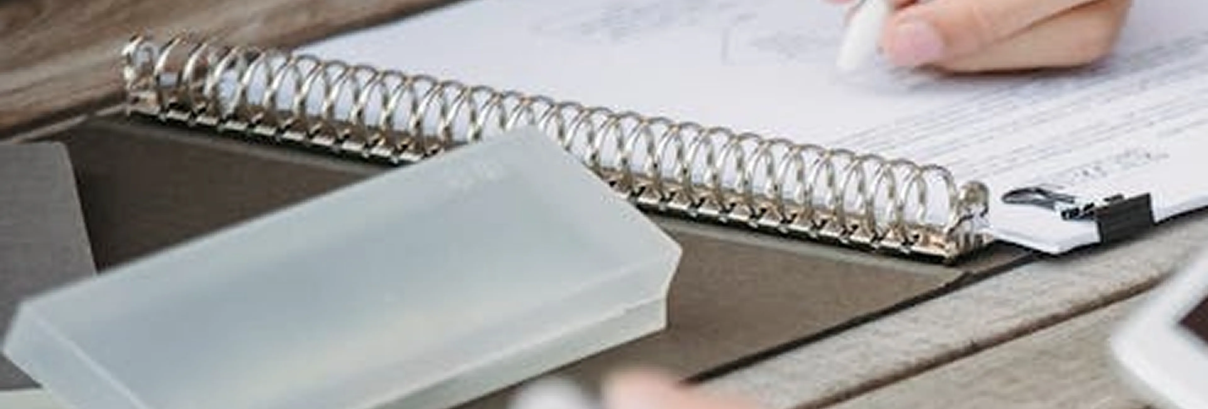 Close-up of a spiral notebook on a wooden table with a translucent plastic case and a hand writing.