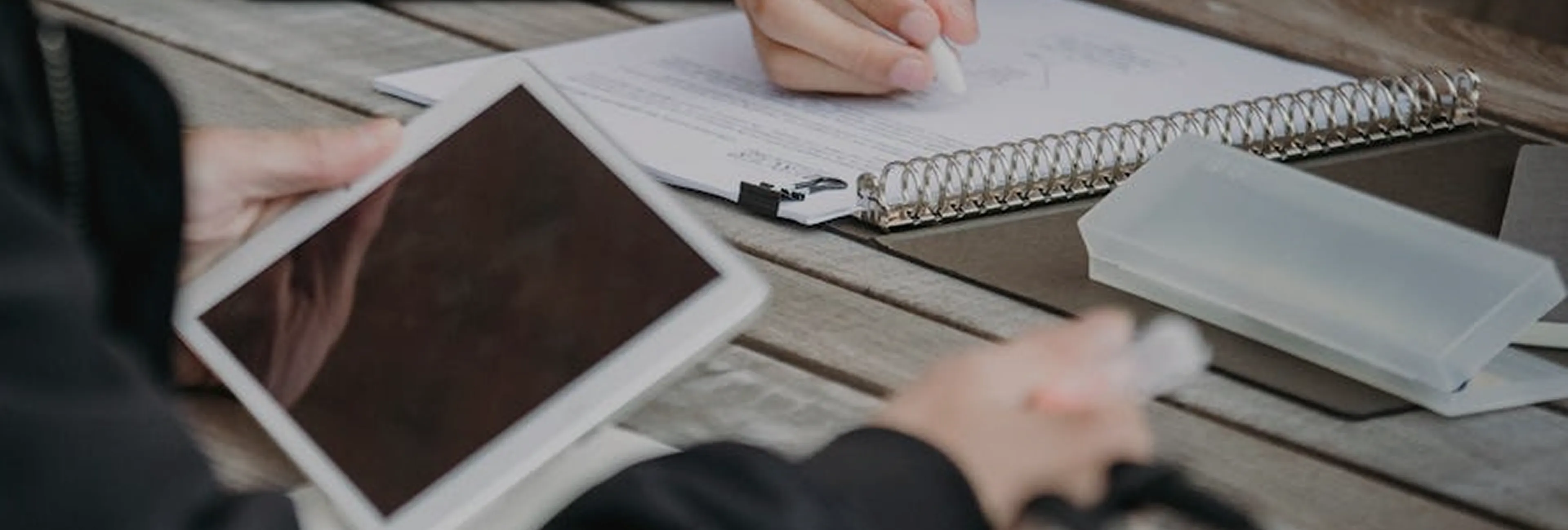Person holding a tablet next to another person writing on a spiral notebook on a wooden table.