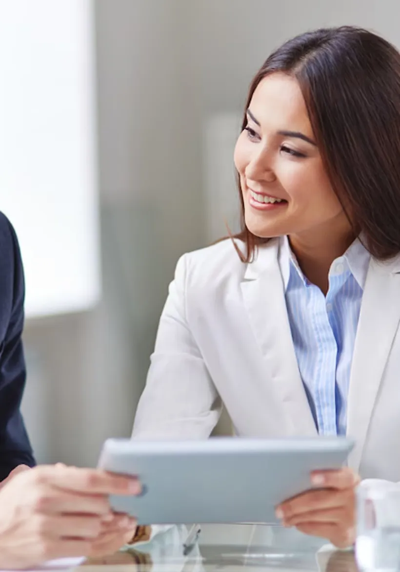 Smiling businesswoman in a white blazer holding a tablet during a discussion.