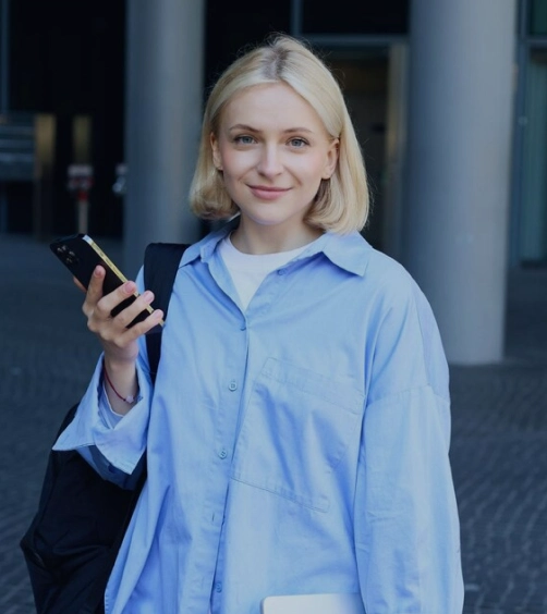 Smiling young woman with blonde hair holding a smartphone, wearing a blue button-up shirt and carrying a black backpack.