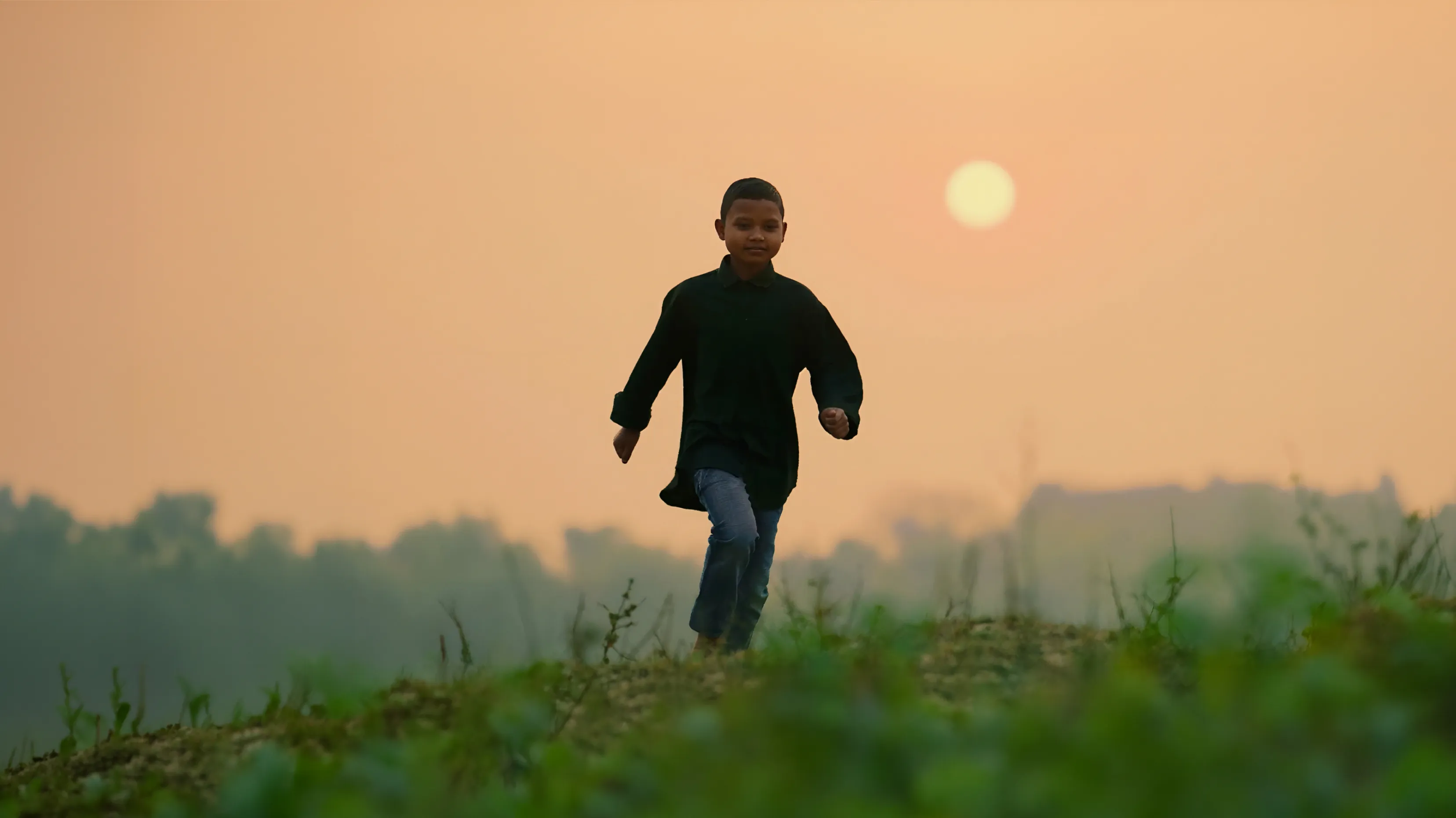 Young boy running on grassy terrain at sunset with an orange sky and distant trees.