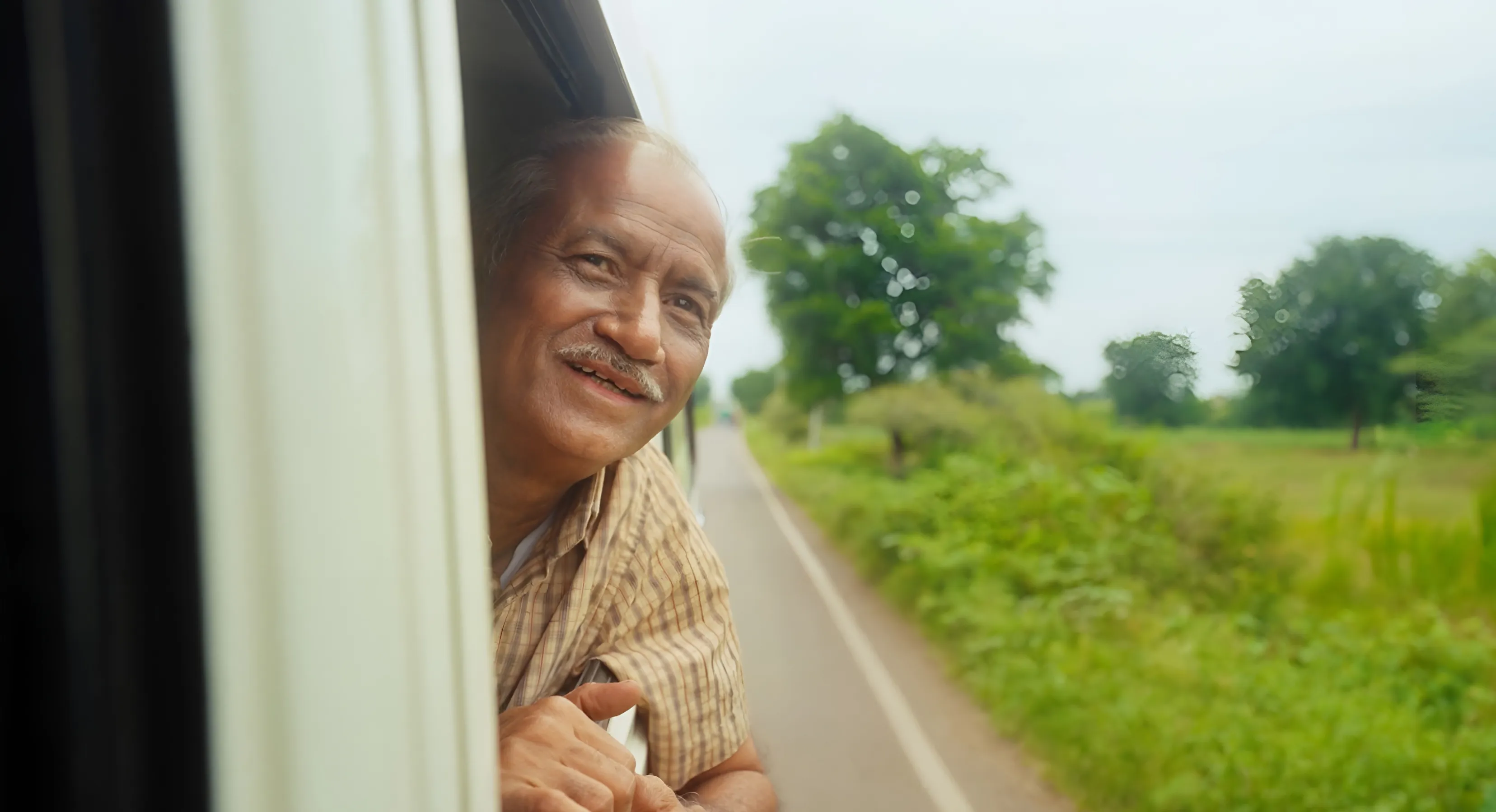 Smiling elderly man leaning out of a vehicle window with a tree-lined road in the background.