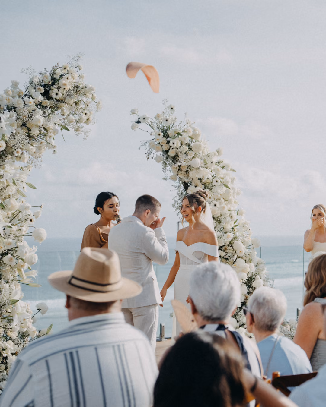 Beachside wedding ceremony with guests and a white floral arch